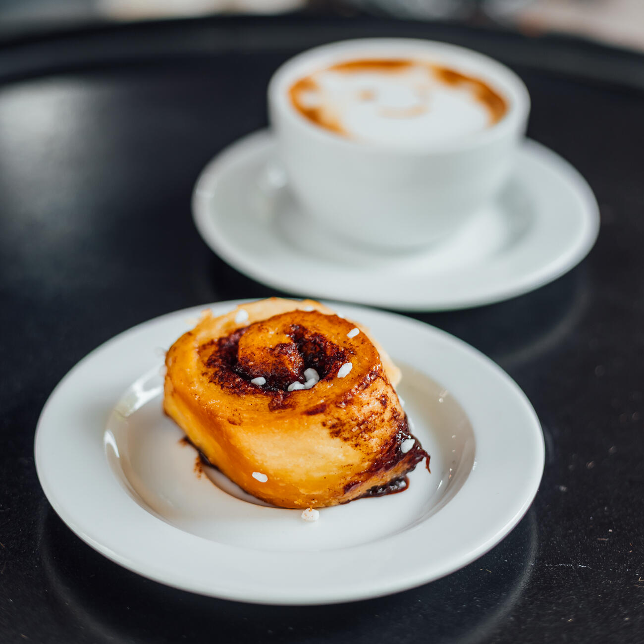 Vegane Zimtschnecke auf weißem Teller mit Cappuccino im Hintergrund – Coffee Deal in der Probierbude am Stephansplatz in Hannover-Südstadt. easy cheese.webp